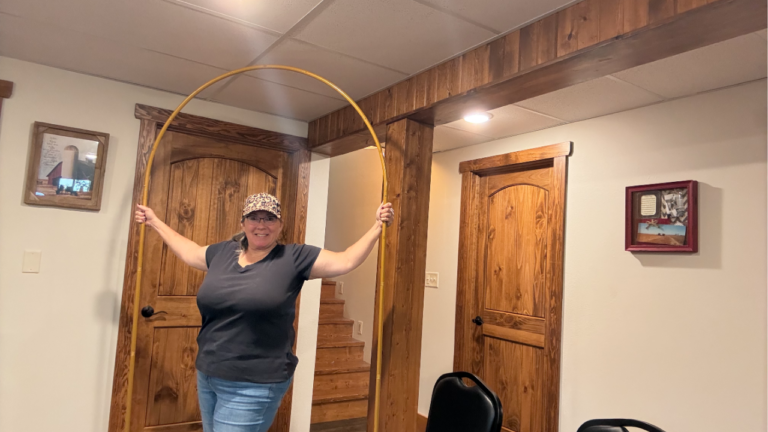 Smiling woman holding a fully assembled gold metal backdrop arch indoors after setup. The decorative arch stands in front of rustic wood doors and demonstrates the size and finished appearance of the Hemmotop metal backdrop stand.